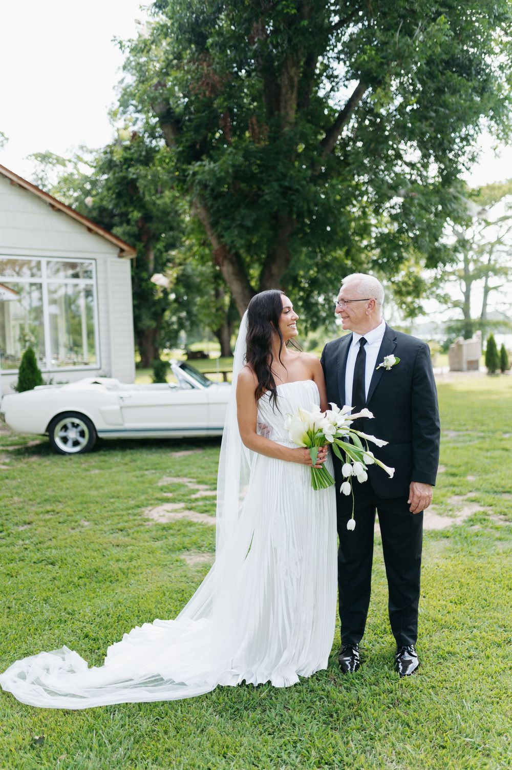 Bride in white strapless gown with long train holds tulip bouquet beside groom in black suit near vintage white convertible.