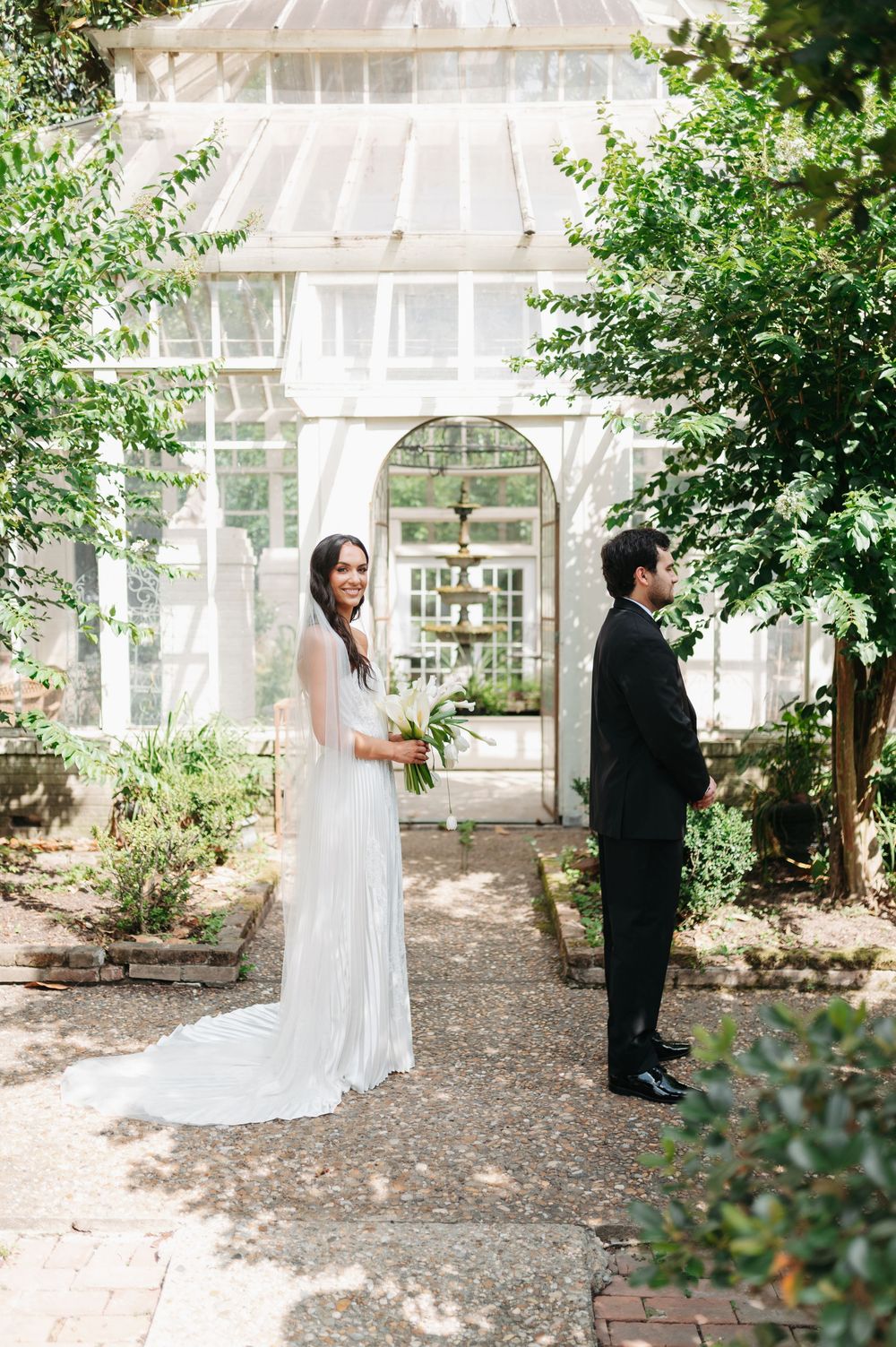 Bride in white gown holds bouquet as groom stands nearby outside a charming greenhouse garden venue.