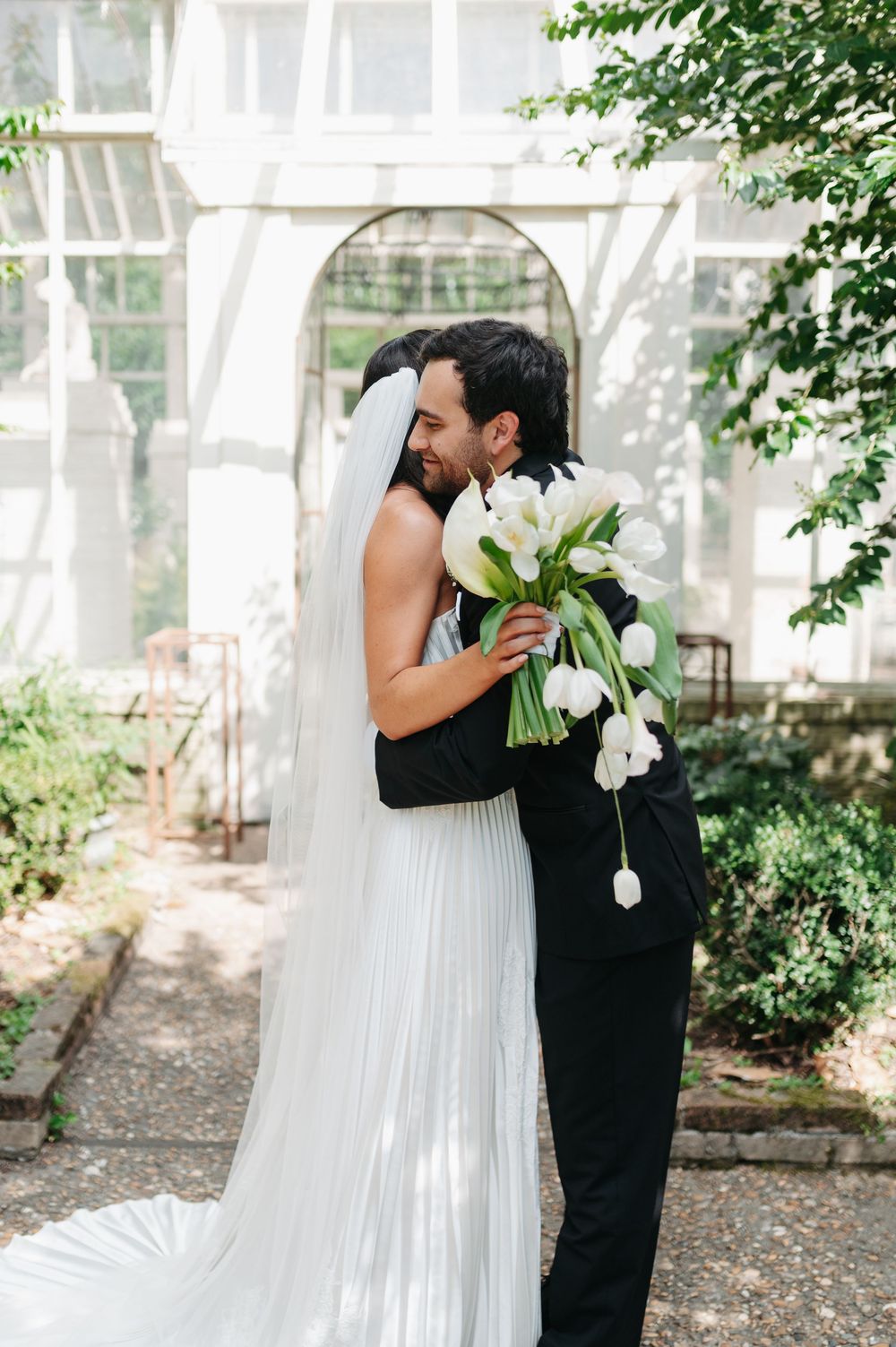 Bride and groom embracing outdoors, bride holding white tulip bouquet with veil trailing, greenhouse arch backdrop.
