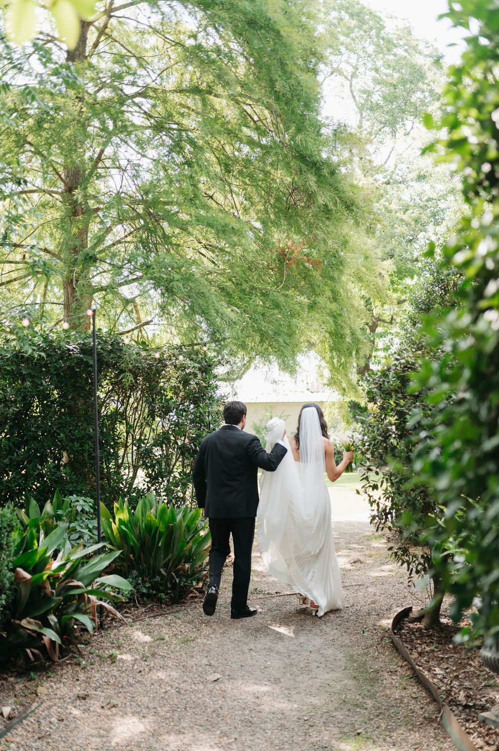 A bride and groom walk hand in hand down a lush garden path lined with green hedges and trees on their wedding day.