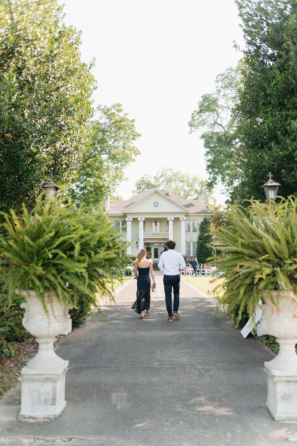 Couple walking hand-in-hand toward an elegant Southern mansion along a tree-lined pathway with ornate stone urns.