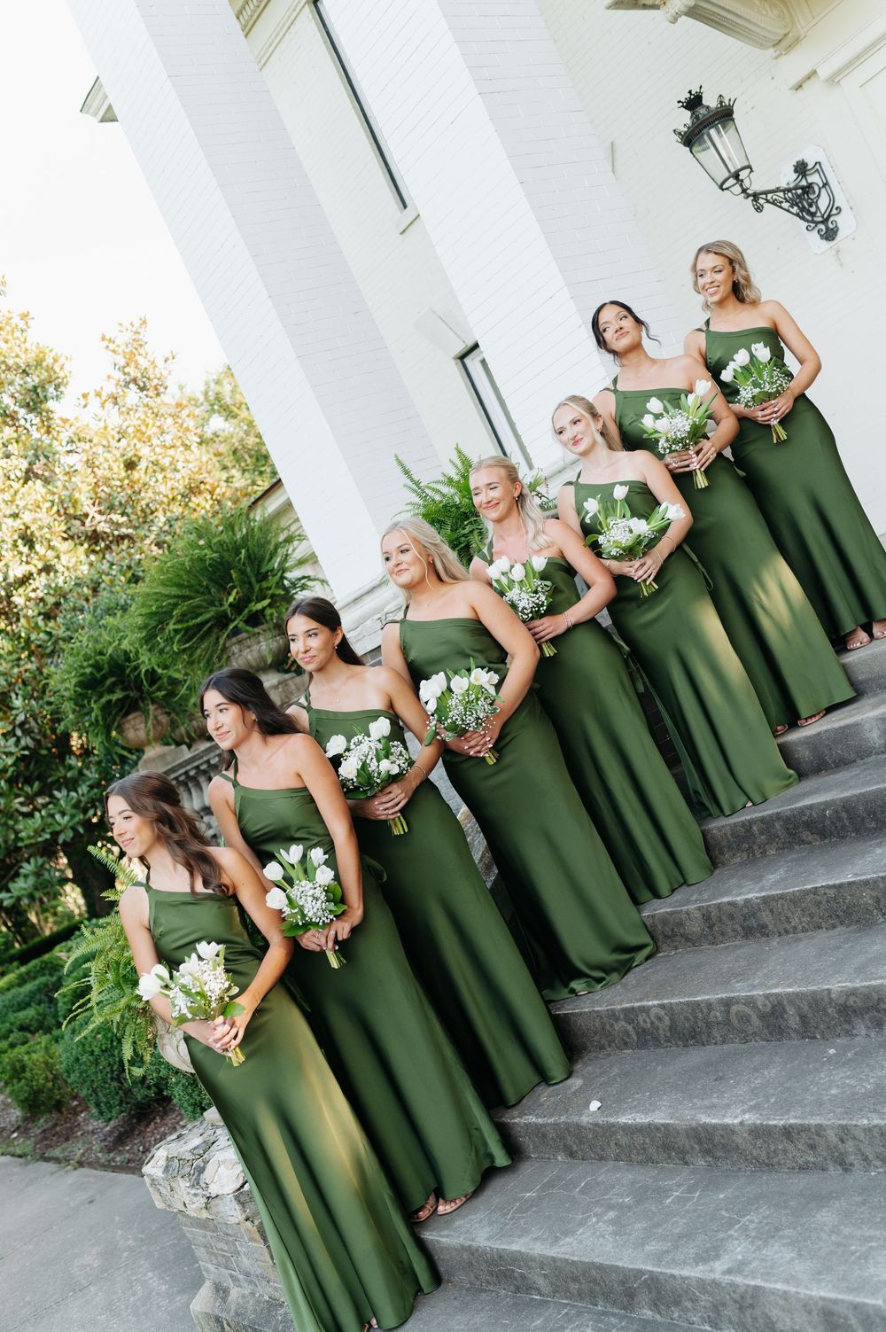 Bridesmaids in olive green gowns holding white bouquets pose on stone steps outside a white columned venue.