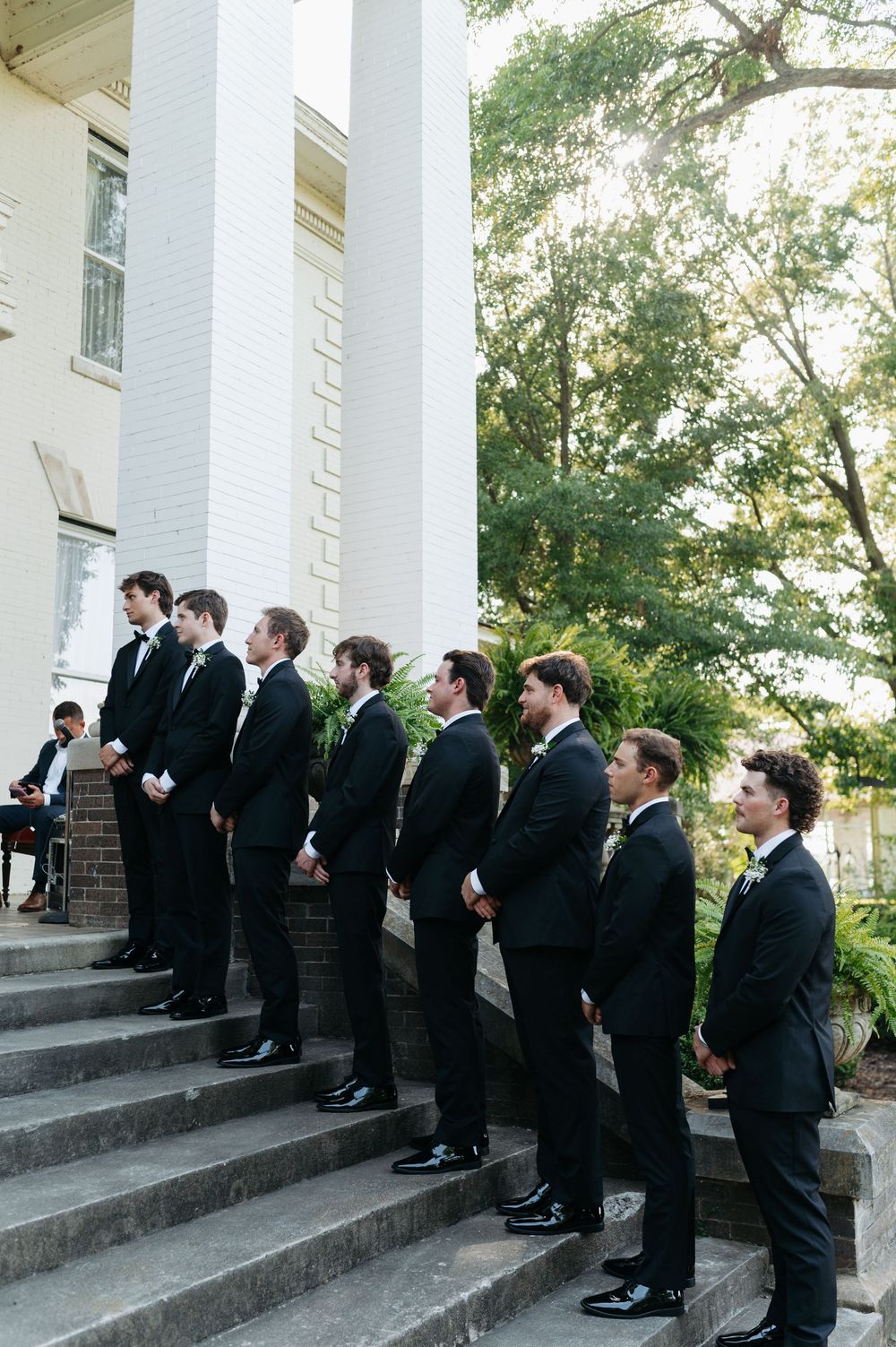 Groomsmen in black tuxedos standing on brick steps outside a white columned building during a wedding.