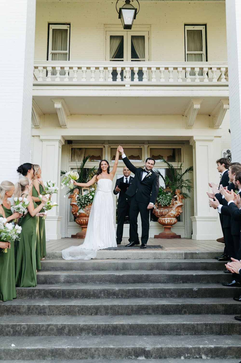 Bride and groom raise hands in celebration on steps of white mansion, flanked by bridesmaids in green dresses.