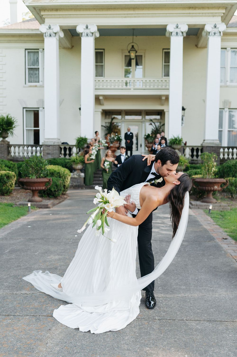Groom dips bride in elegant white gown for romantic kiss outside grand Southern mansion wedding venue.