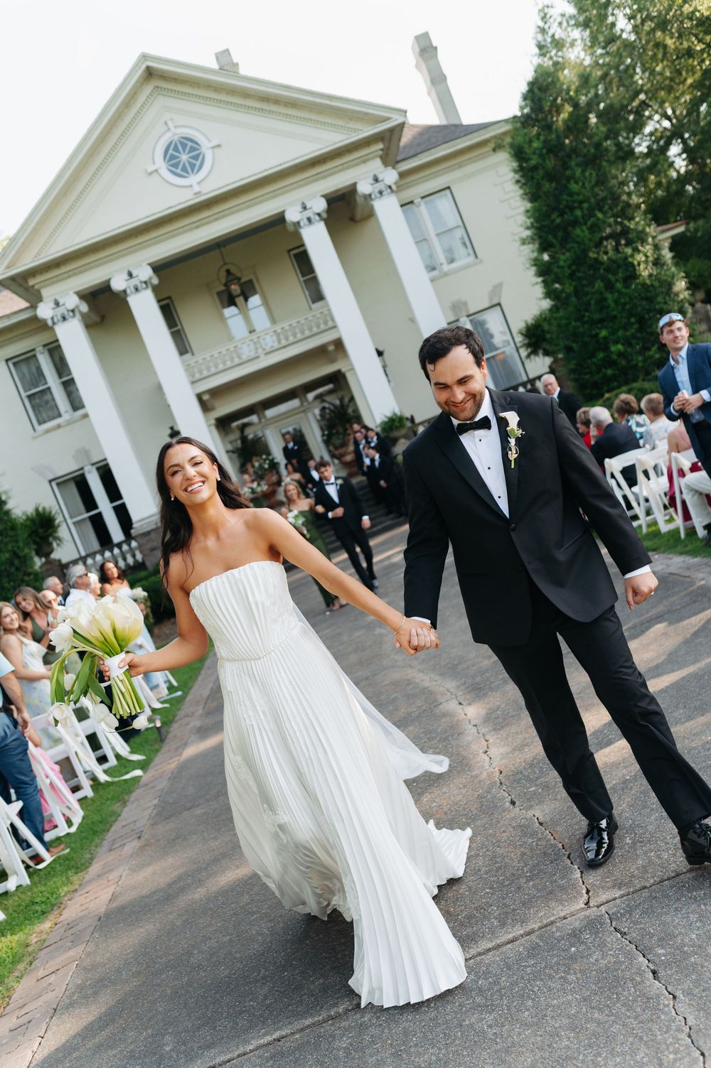 Joyful newlyweds hold hands walking down the aisle after outdoor wedding ceremony at elegant white colonial mansion.