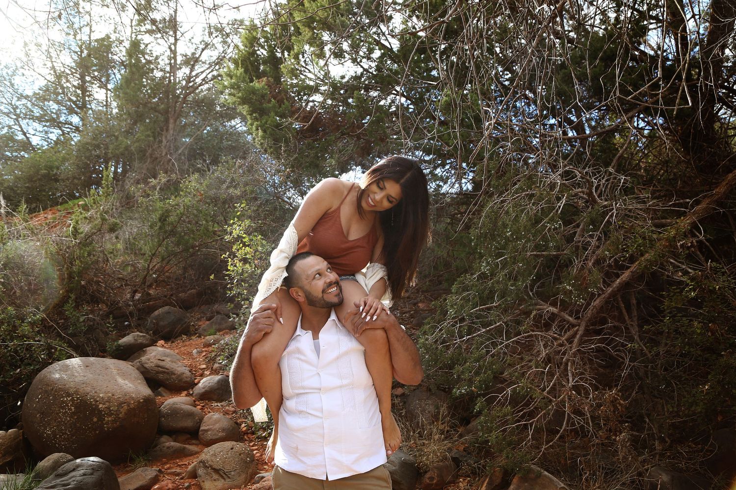 Smiling woman sits on a man's shoulders among rocks and trees in a sunlit outdoor couple's portrait.