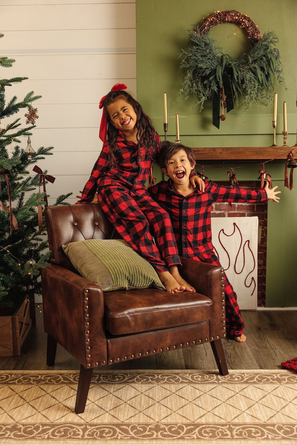 Two kids in matching red plaid Christmas pajamas laugh joyfully on a leather chair by a decorated tree and fireplace.