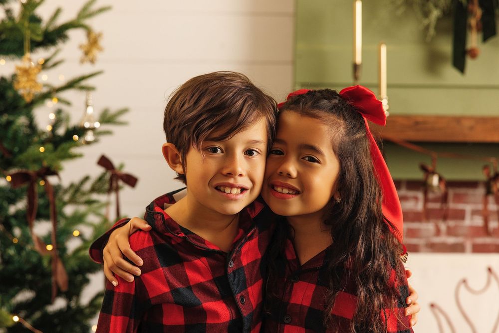 Two children in matching red plaid pajamas smile together near a decorated Christmas tree in a festive holiday setting.