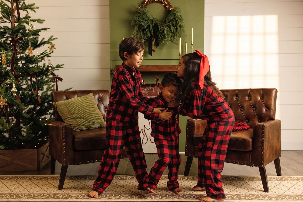 Three children in matching red buffalo plaid Christmas pajamas playing together near a decorated holiday tree.
