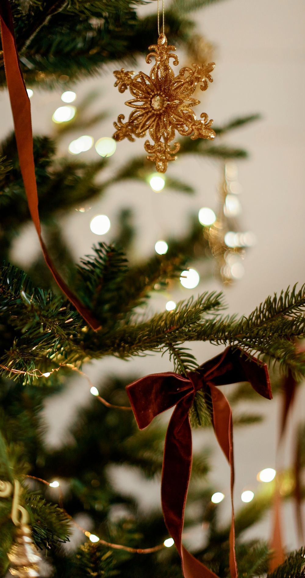 Gold glitter snowflake ornament and red velvet bow on a Christmas tree with warm bokeh lights in background.