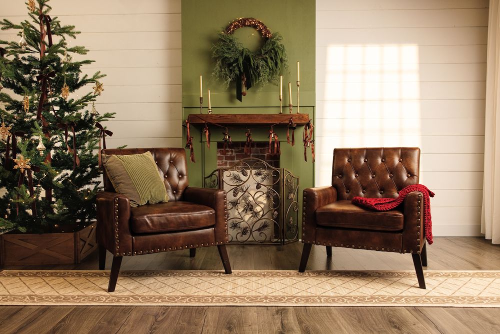 Two brown leather tufted armchairs beside a Christmas tree and fireplace with wreath in a festive holiday living room.