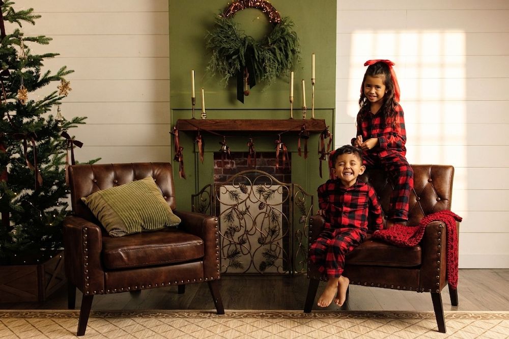 Two children in matching red plaid Christmas pajamas pose by a decorated fireplace and tree in a cozy holiday living room.