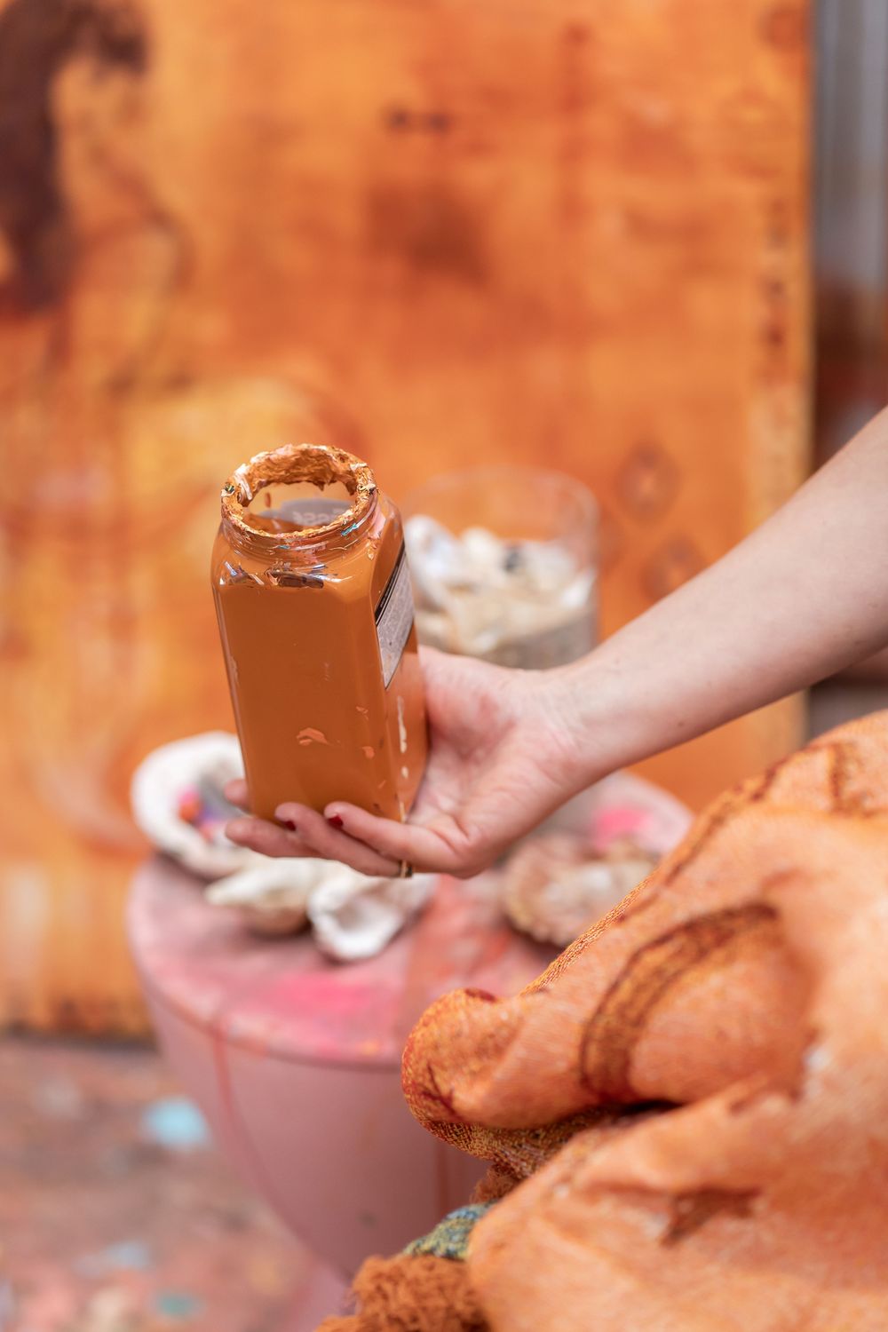 Hand holding a glass jar of brown paint in an artist's studio with palette and orange canvas in background.