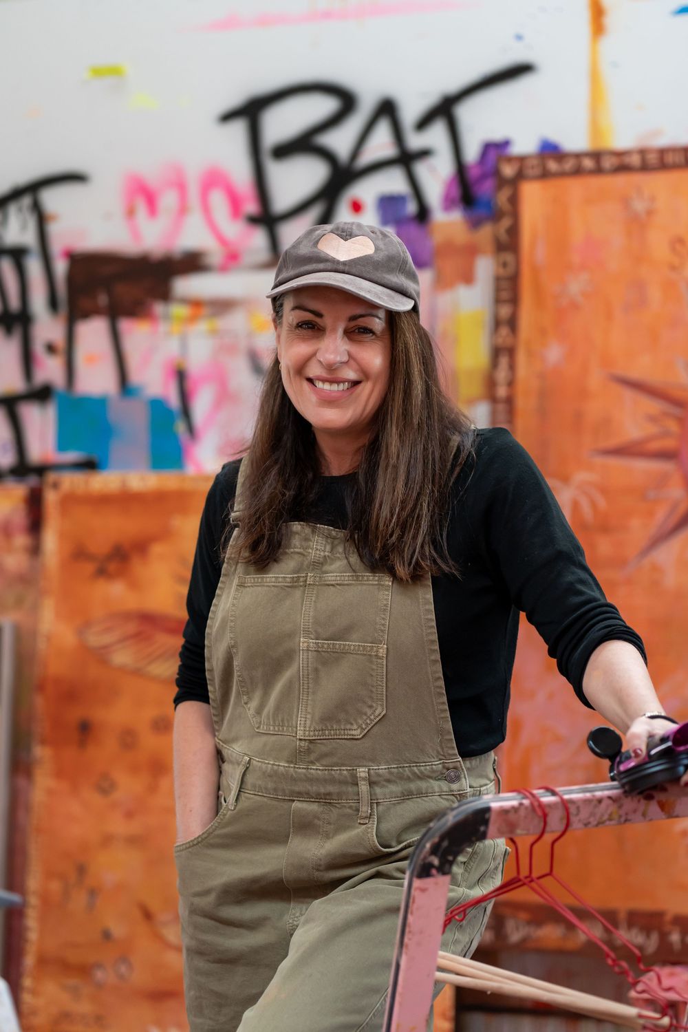 Smiling woman in olive overalls and heart cap poses in front of colorful graffiti wall in art studio.