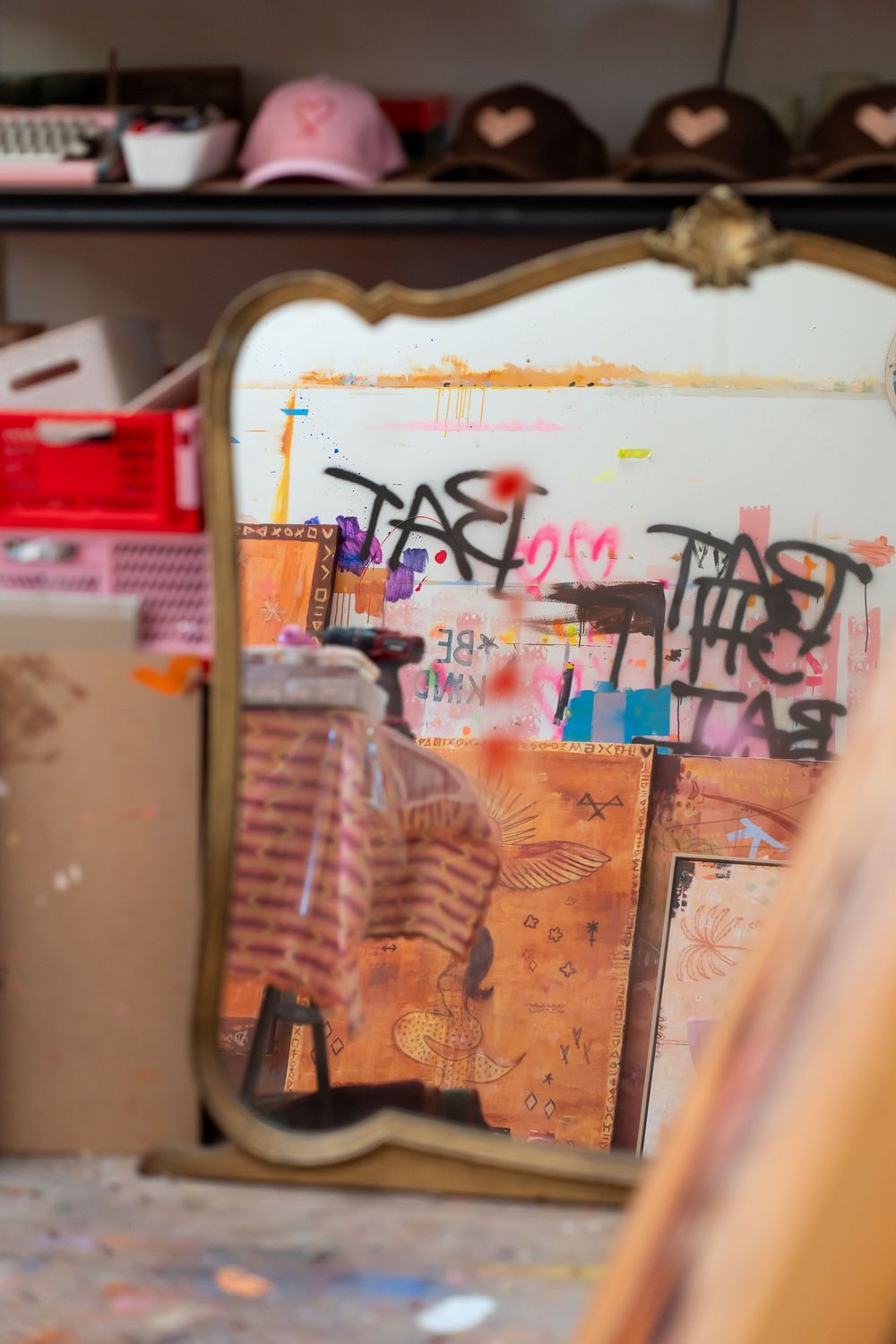 Vintage gold-framed mirror reflecting graffiti art and paintings in a cluttered artist's studio with hats on shelves.