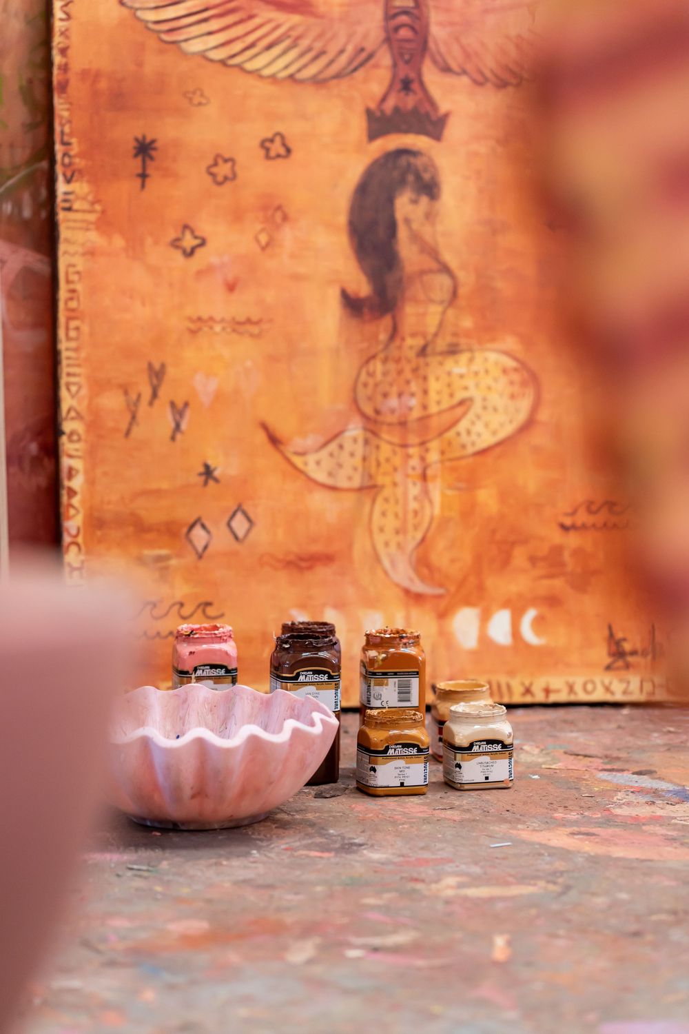 Artist's studio with paint jars on floor before an orange Egyptian-inspired mural featuring a goddess figure.