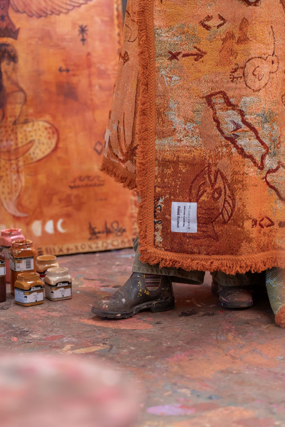 Person wearing boots draped in an orange patterned rug with fringe, surrounded by paint jars in a colorful studio.
