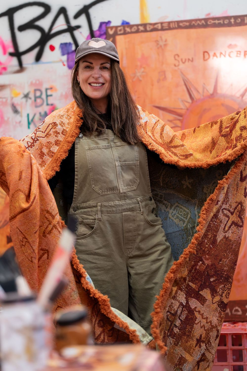 Woman in green overalls and cap holds orange patterned textile in front of colorful graffiti wall art.