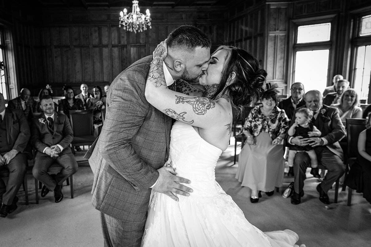 Bride and groom share their first kiss at wedding ceremony in elegant wood-paneled venue with guests watching.