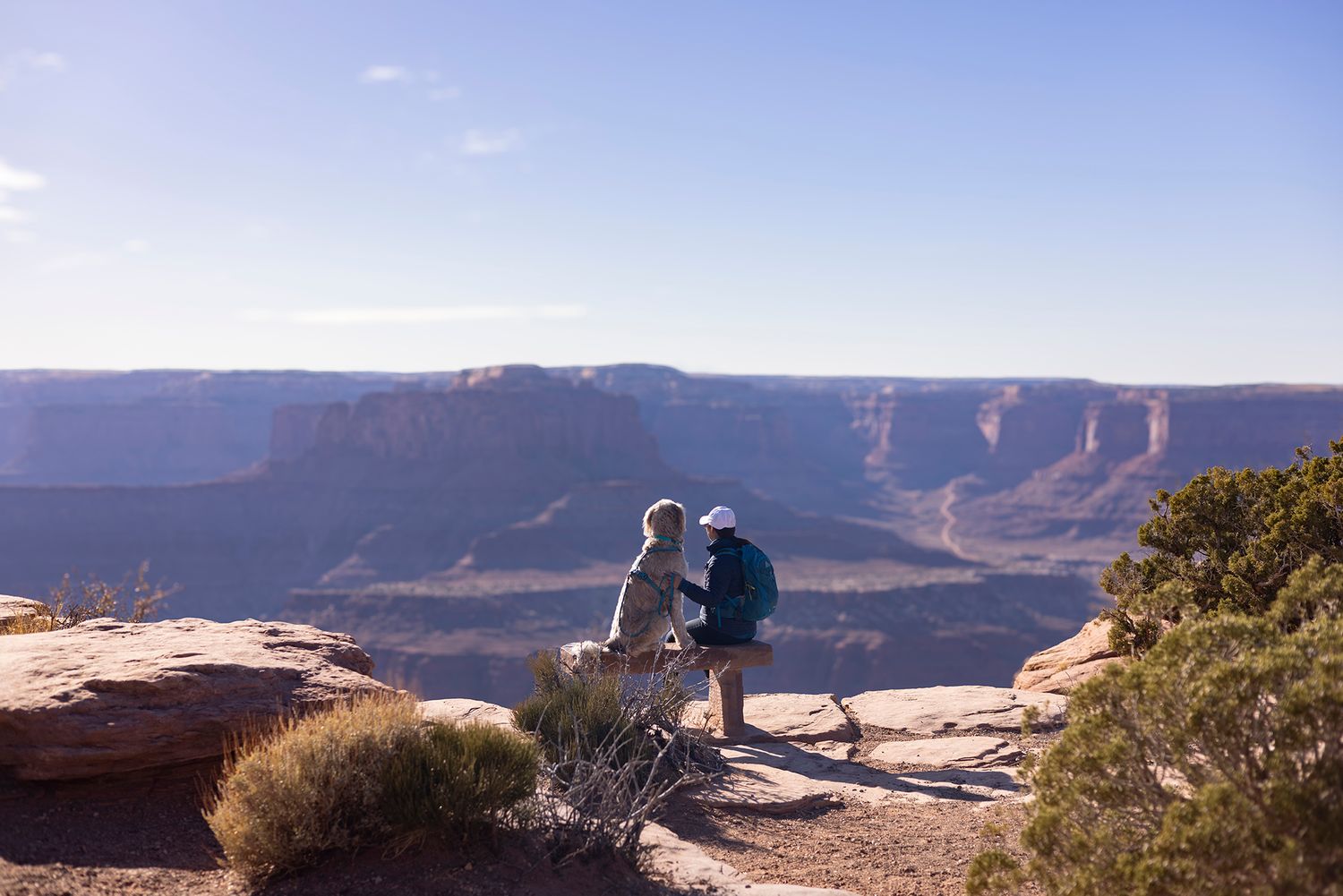 Two hikers stand on a rocky overlook admiring the vast canyon landscape under a clear blue sky.