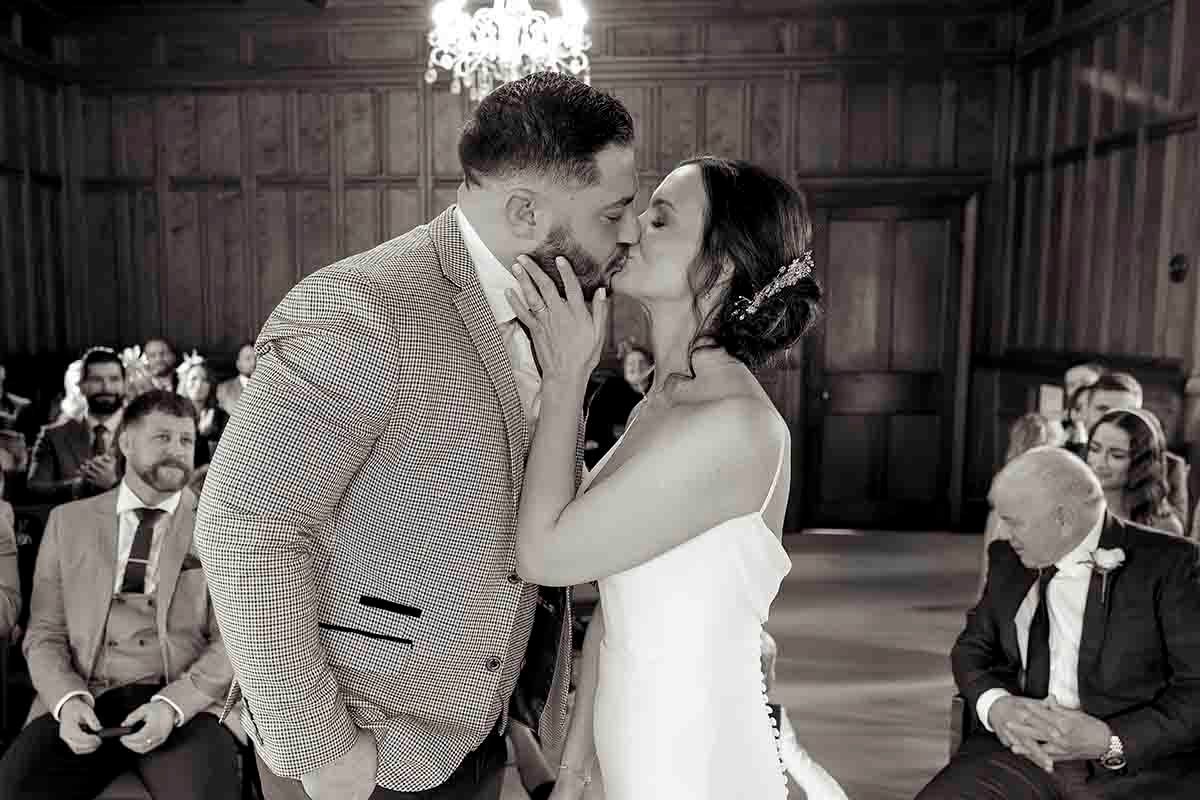 Black and white photo of a couple sharing their first kiss at a wedding ceremony with guests seated in the background.