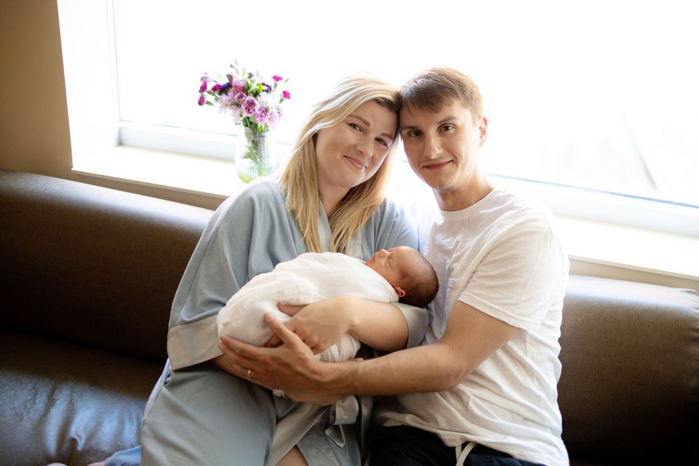 Young couple sitting on a leather sofa, smiling and holding their newborn baby wrapped in white University of Tennessee Medical Center.