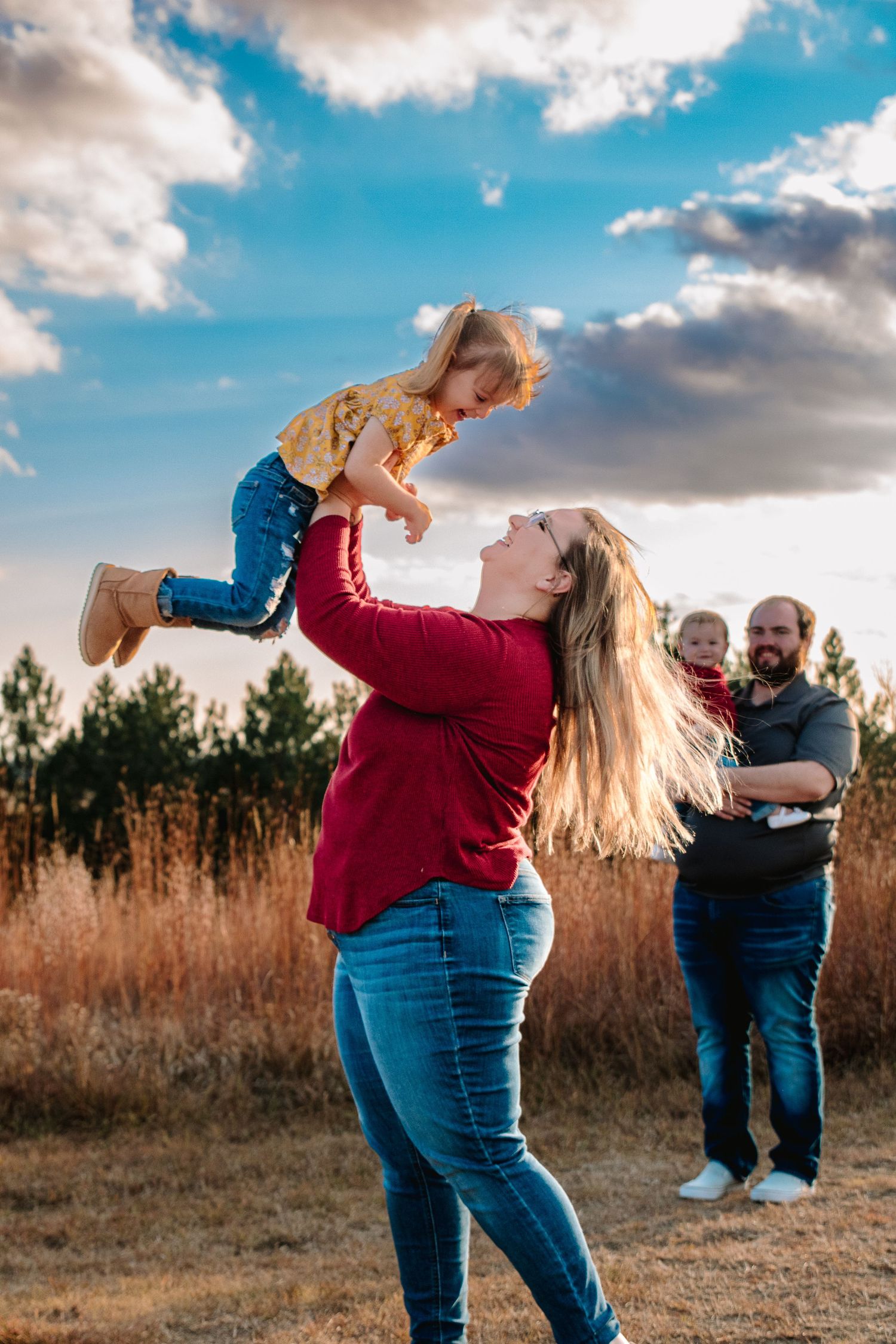 A joyful mother lifts her toddler into the sky while father holds baby in a sunlit autumn field.