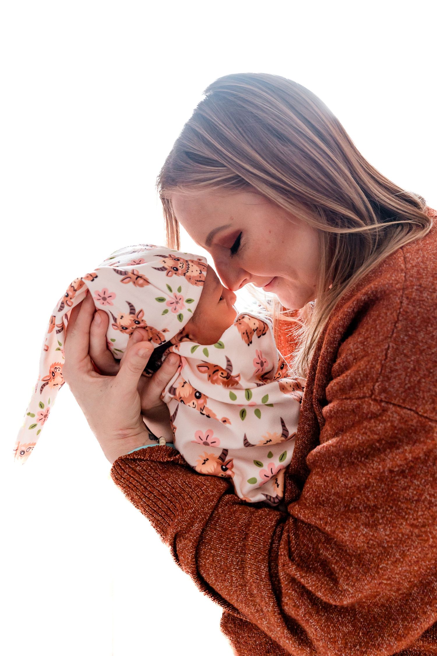 A smiling mother in a rust sweater tenderly holds her newborn baby dressed in a floral fox outfit and knotted hat.