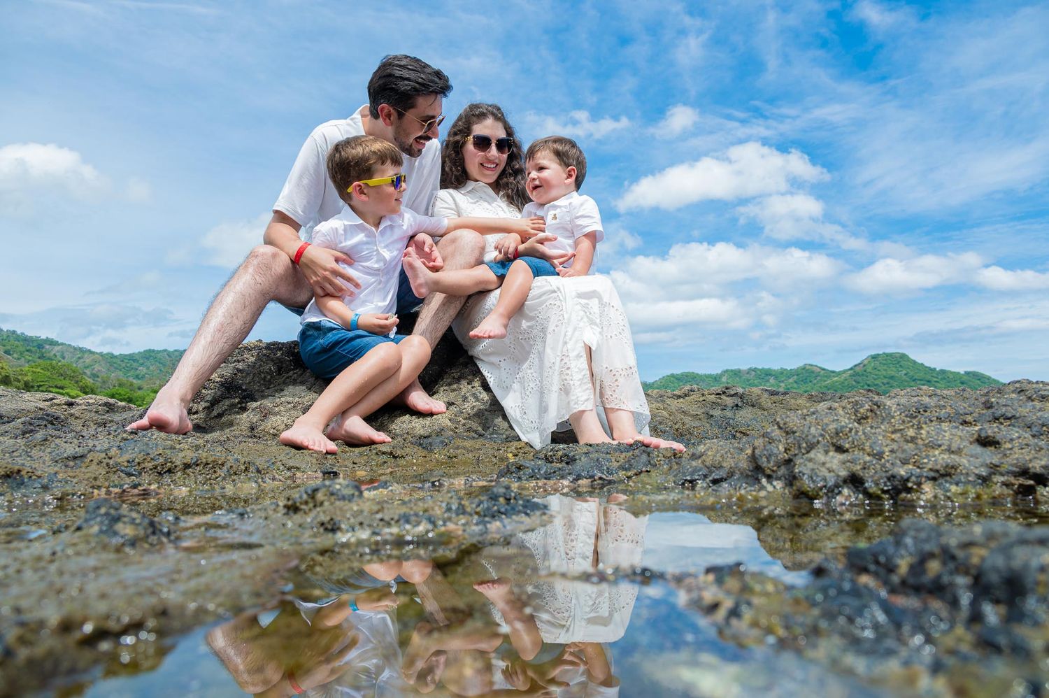 Family of four sitting on rocky shoreline with green hills and blue sky, reflections visible in tidal pool.