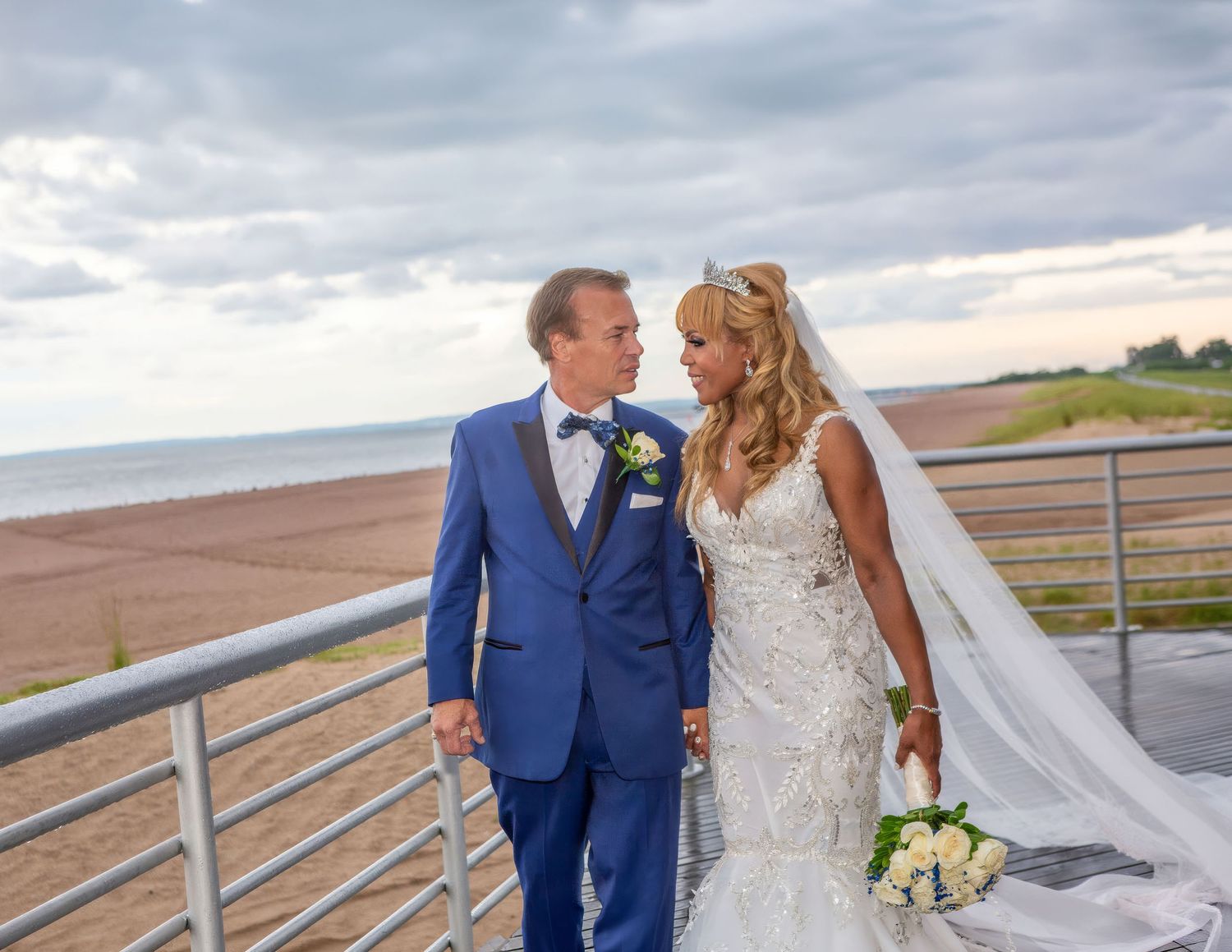 A bride and groom share a romantic moment on a seaside deck, she in a lace gown with veil, he in a blue suit.