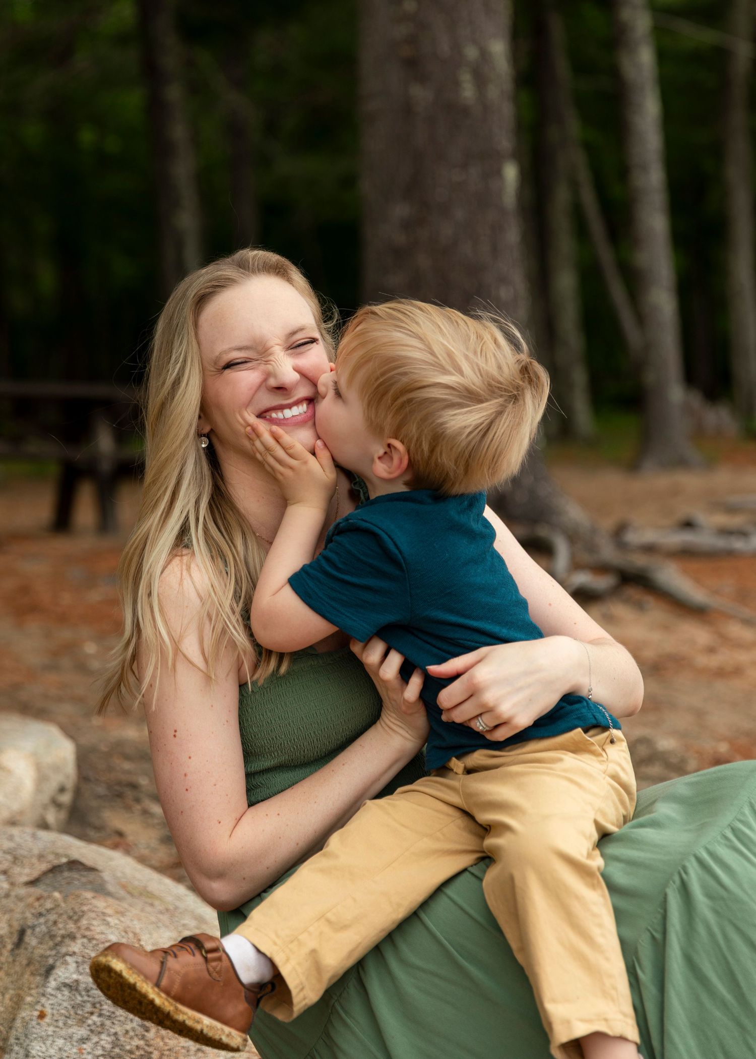 A joyful blonde mother laughs as her young toddler son gives her a sweet kiss on the cheek outdoors.