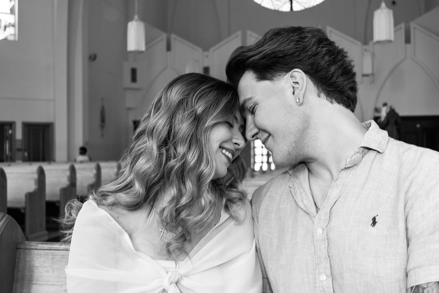 Black and white image of a couple sitting in a church, foreheads touching and smiling with their eyes closed