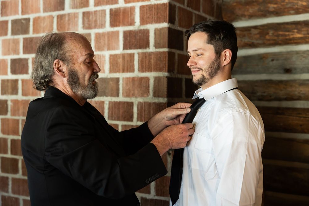 Older man helping young groom adjust bow tie before wedding ceremony near rustic brick wall.