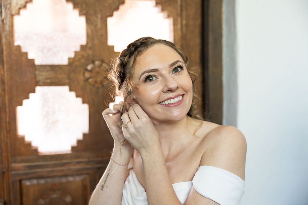 Smiling bride adjusting earring, wearing white dress, standing near ornate wooden door.