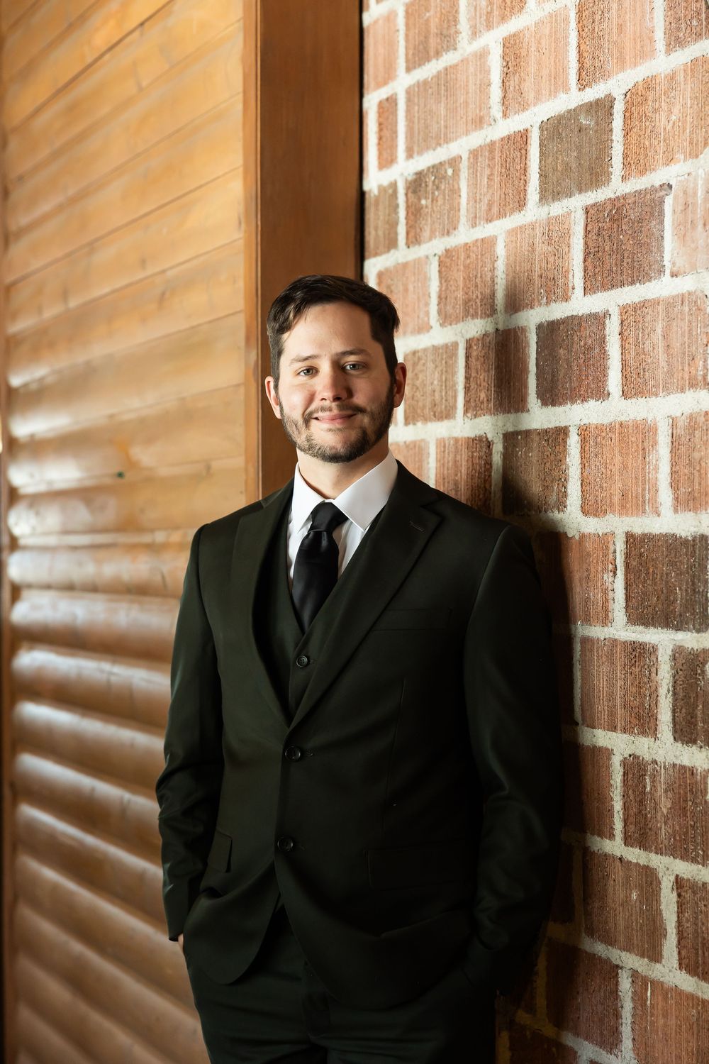 A confident young man in a dark suit and tie smiles while leaning against a brick wall indoors.
