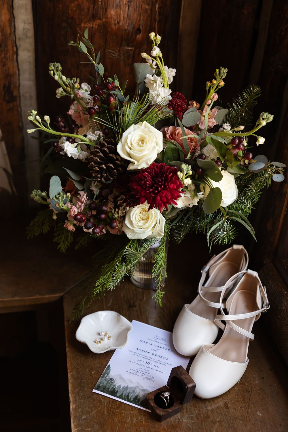 Winter wedding flat lay with white bridal shoes, bouquet of red dahlias, white roses, pinecones, and wedding invitation on wooden surface.