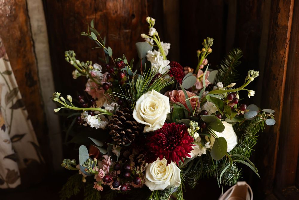Elegant winter wedding bouquet with white roses, dark red dahlias, pine cones, eucalyptus, and berries on rustic wood.
