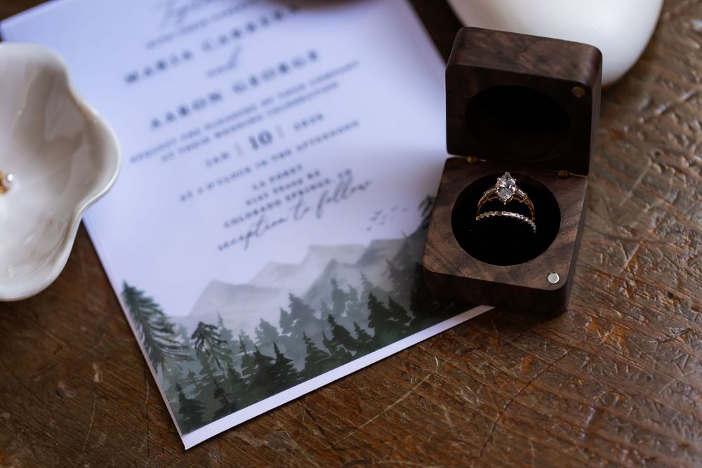 Elegant wedding invitation with mountain forest design beside a diamond engagement ring in a wooden ring box.