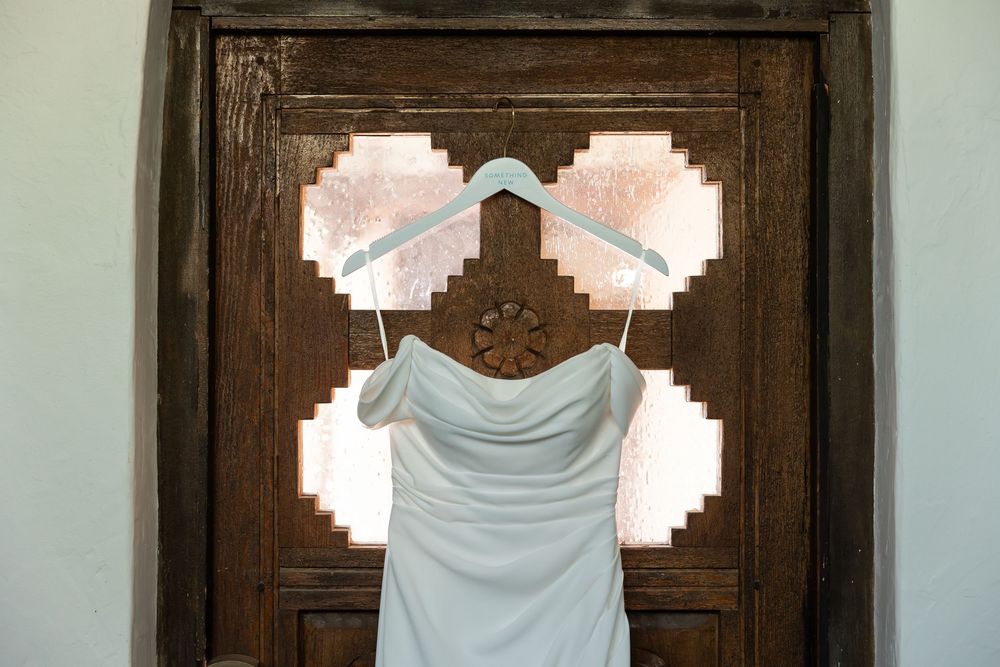 Elegant white wedding dress hanging on mint hangers against a rustic wooden door with decorative stained glass panels.
