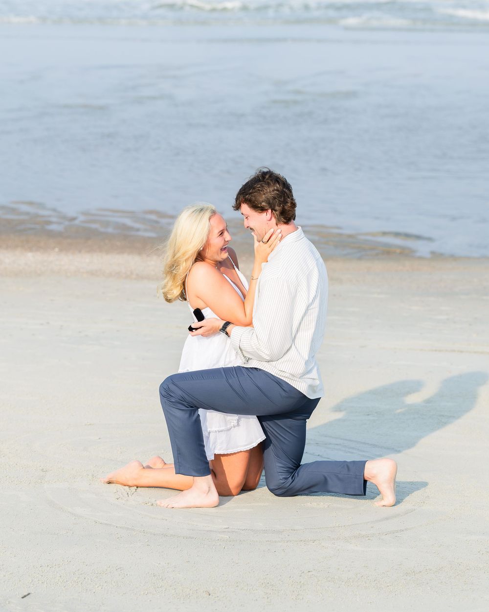Couple sharing a romantic beach proposal moment, man on one knee holding ring box, woman smiling joyfully by ocean shore.