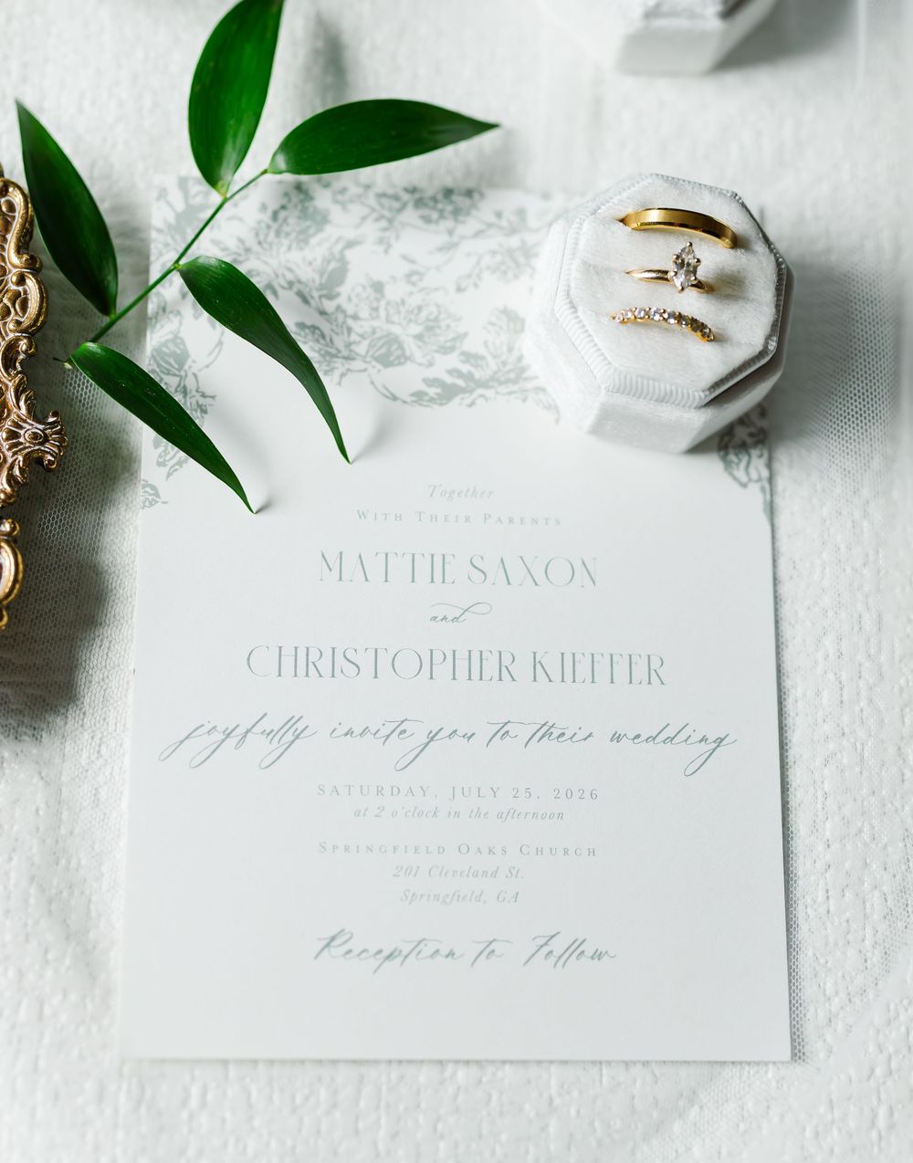 Elegant wedding invitation for Mattie Saxon and Christopher Kiefer with gold rings in a white velvet box and green botanical accents.