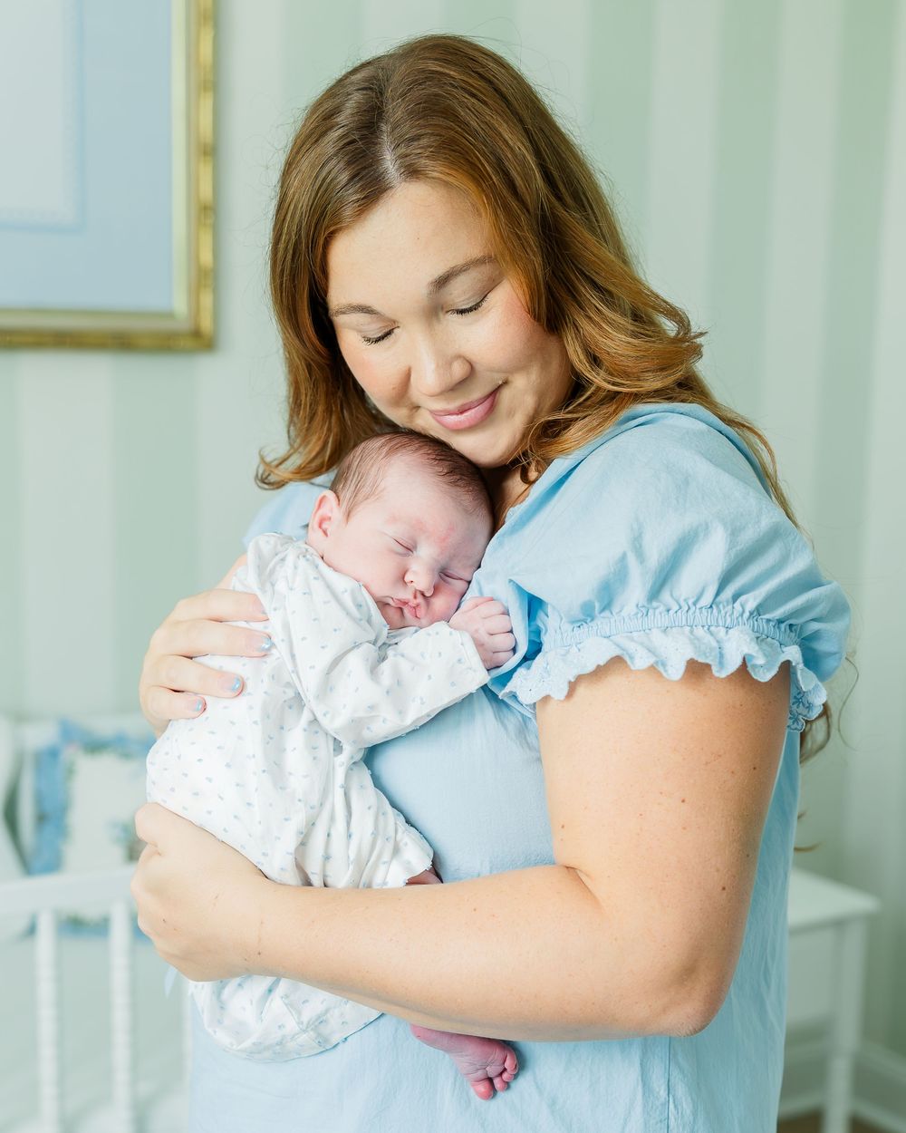 A smiling mother in a blue dress tenderly holds her sleeping newborn baby in a softly lit nursery.
