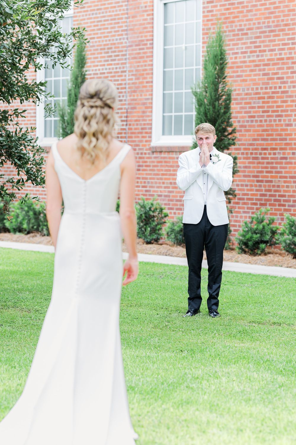Groom in white tuxedo jacket clasps hands in awe during first look with bride in elegant white gown outside brick church.