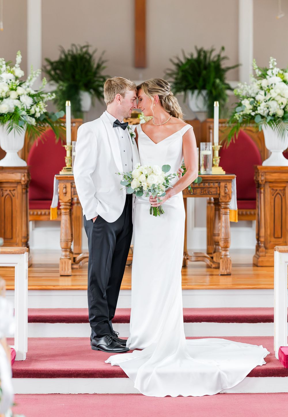 Bride and groom share a romantic moment at the church altar, she holds a white bouquet in an elegant satin gown.