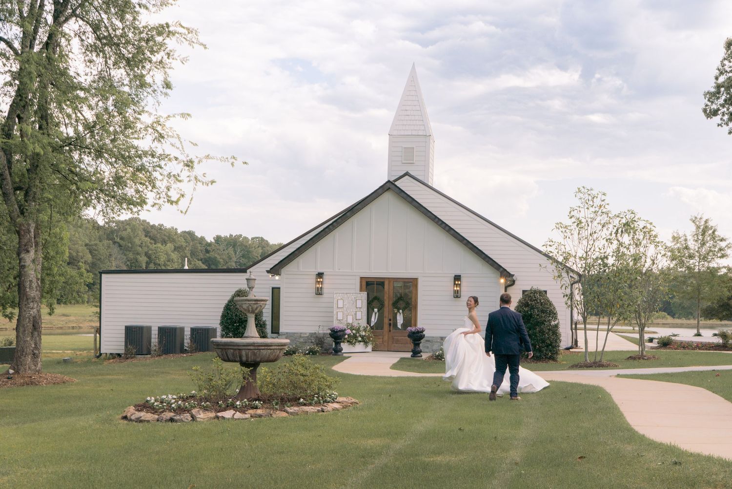 Bride and groom walking toward a white chapel wedding venue with steeple, fountain, and lush green grounds.