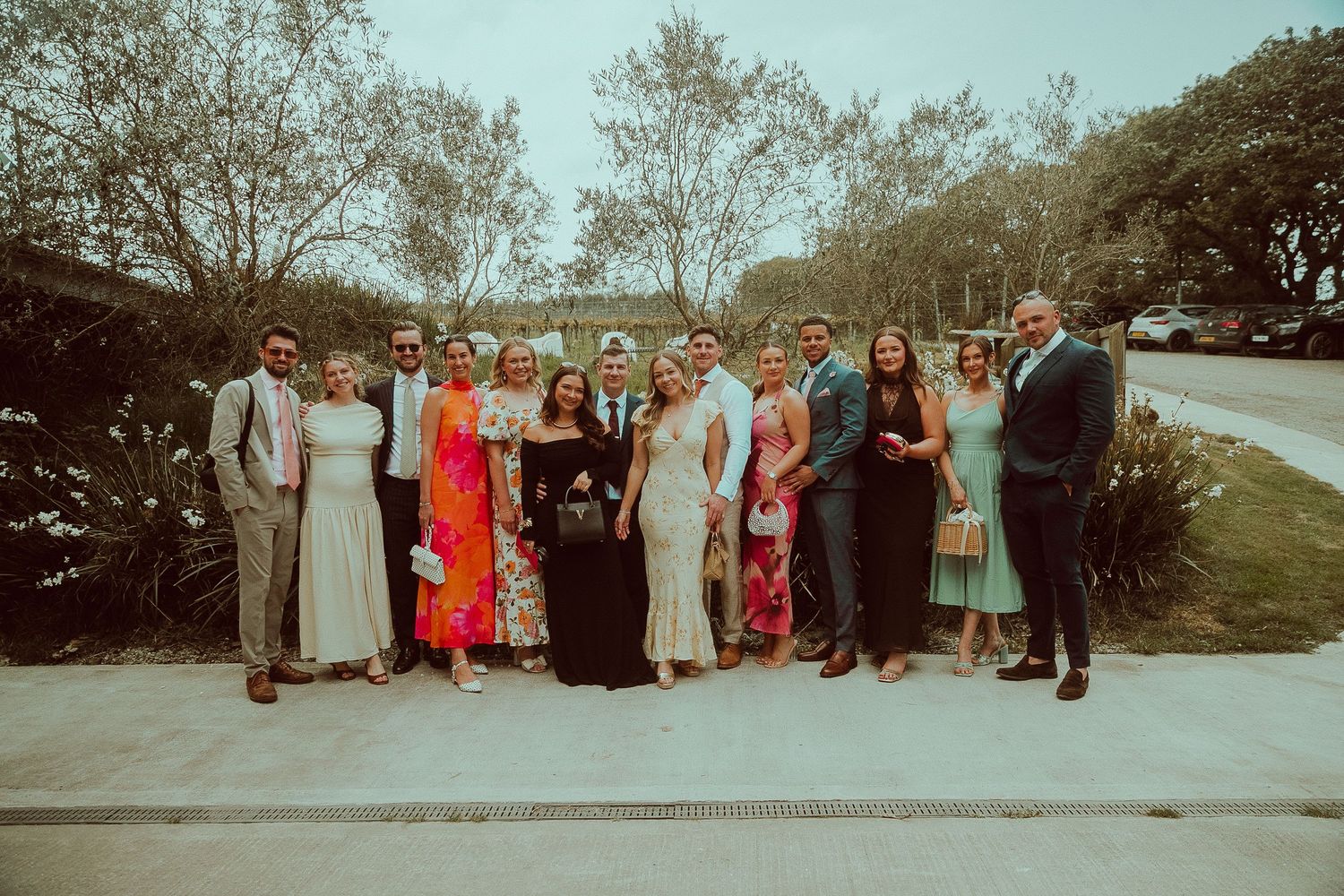 Large group of elegantly dressed wedding guests posing together outdoors on a sunny day.