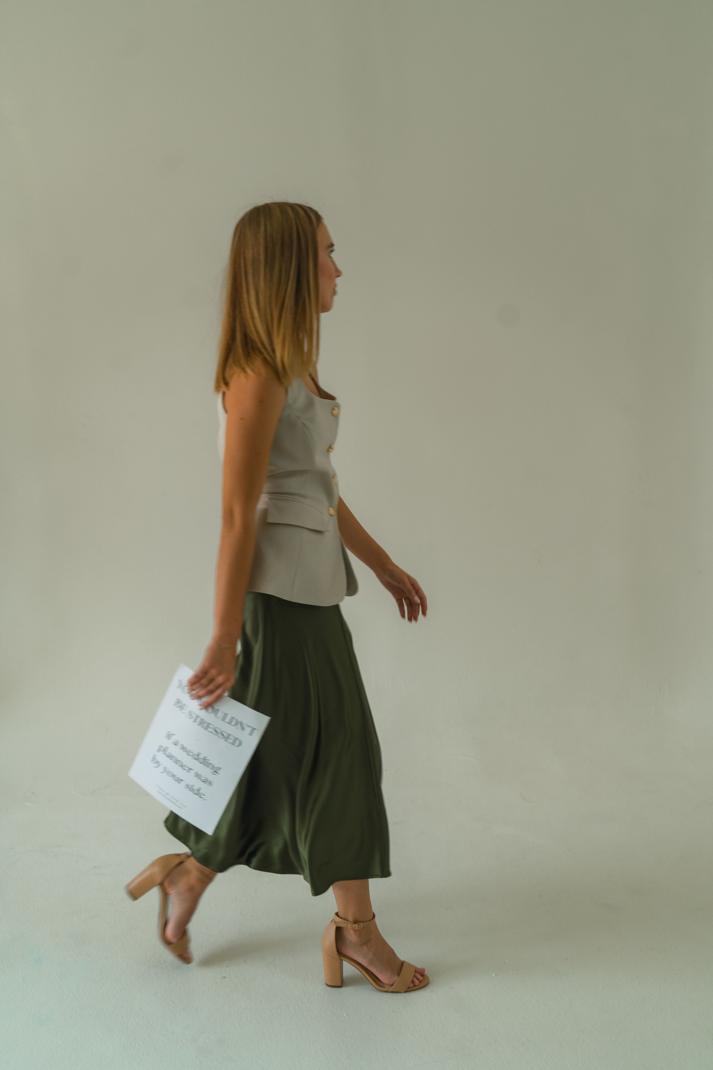 Woman walking in olive green midi skirt, beige vest top, and tan heeled sandals, carrying white papers.
