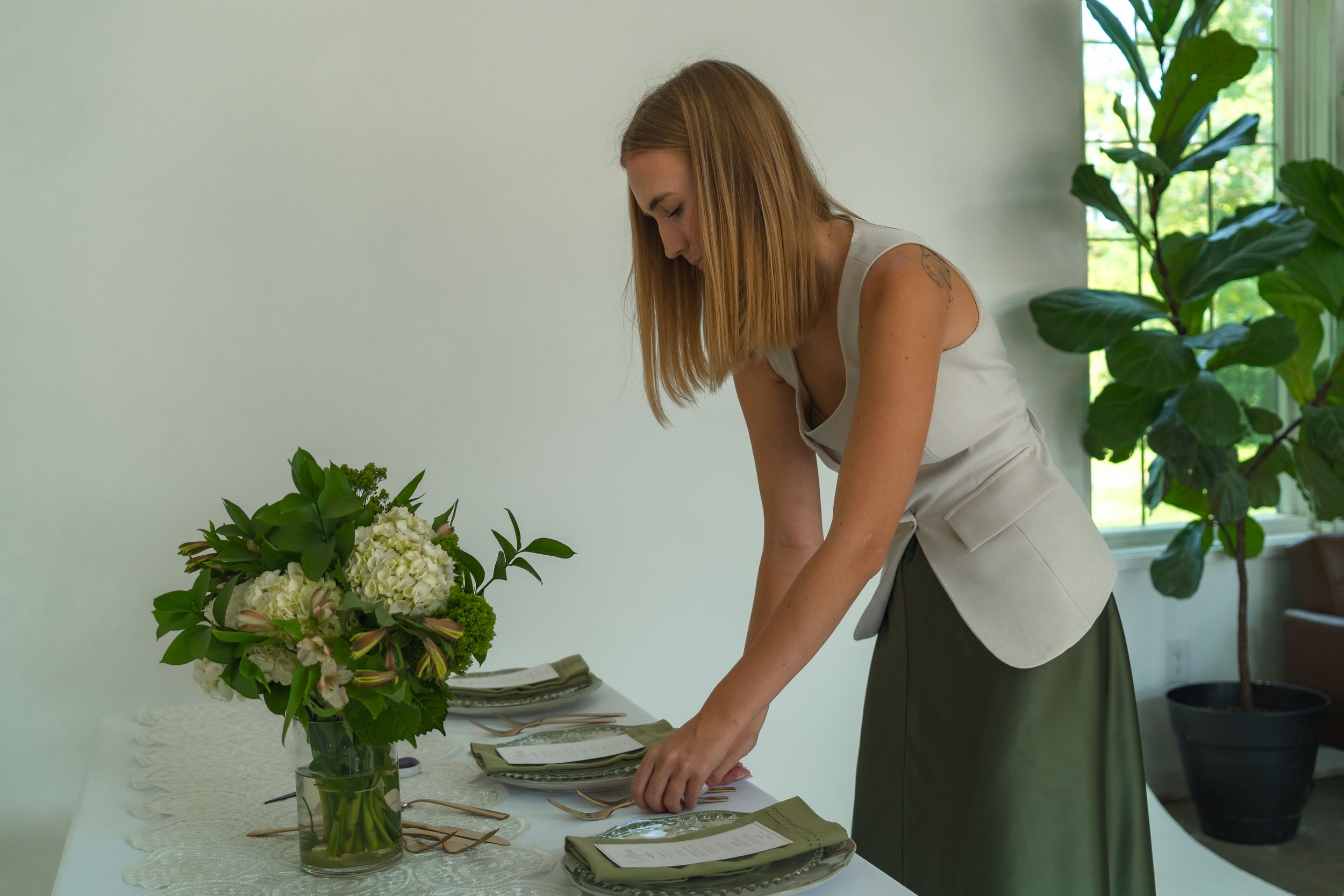 Woman arranging napkins on a white tablecloth beside a green hydrangea centerpiece in a bright minimalist room.