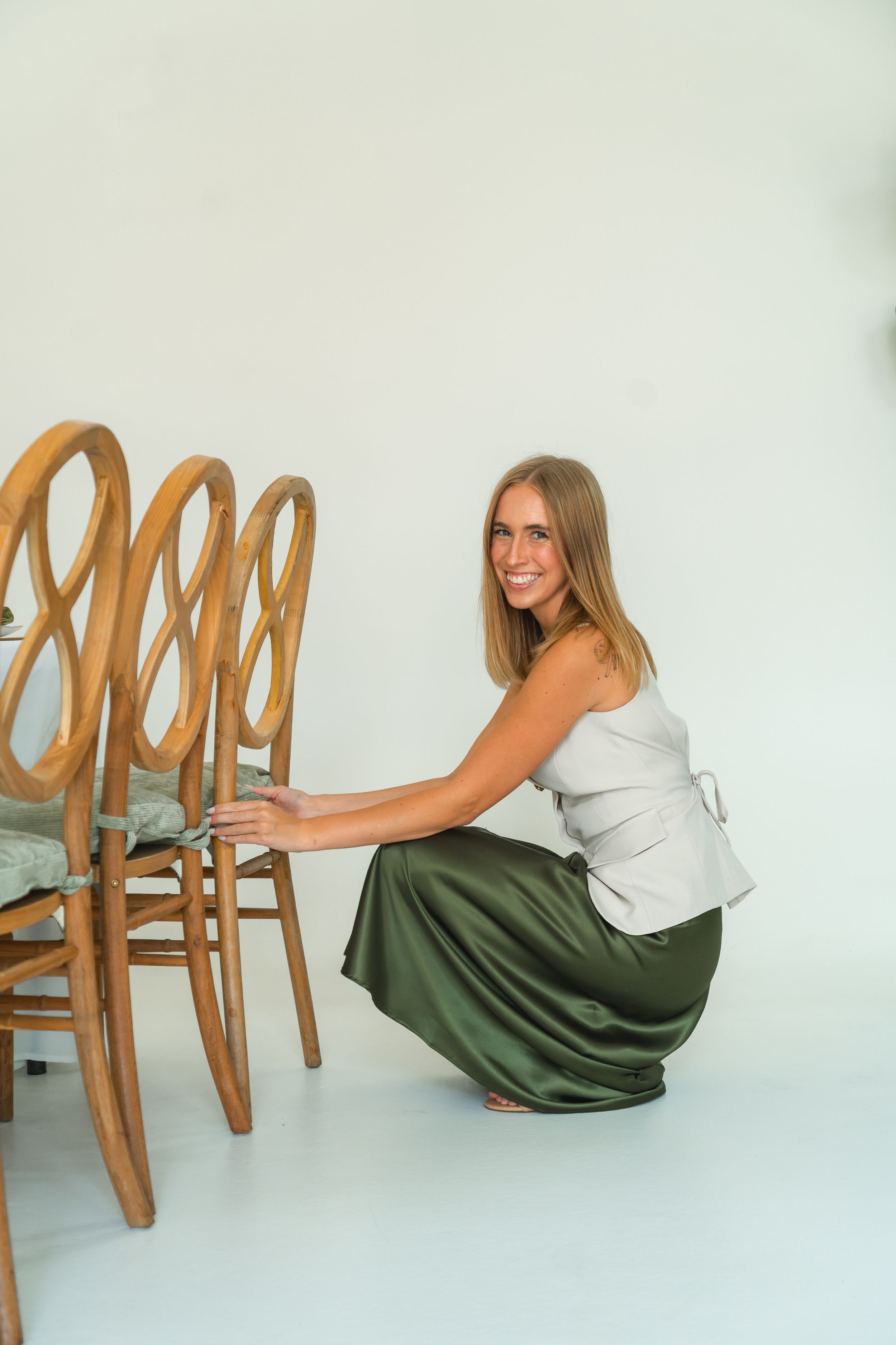 Smiling woman in green satin skirt and white top crouching beside wooden bentwood chairs against white background.